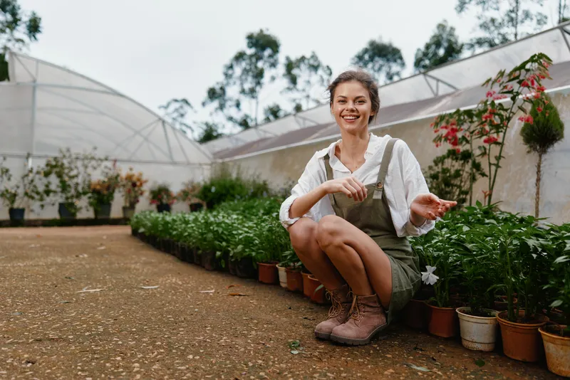 Happy young woman in overalls gardening, surrounded by plants in a greenhouse, showcasing her love for nature and sustainable living