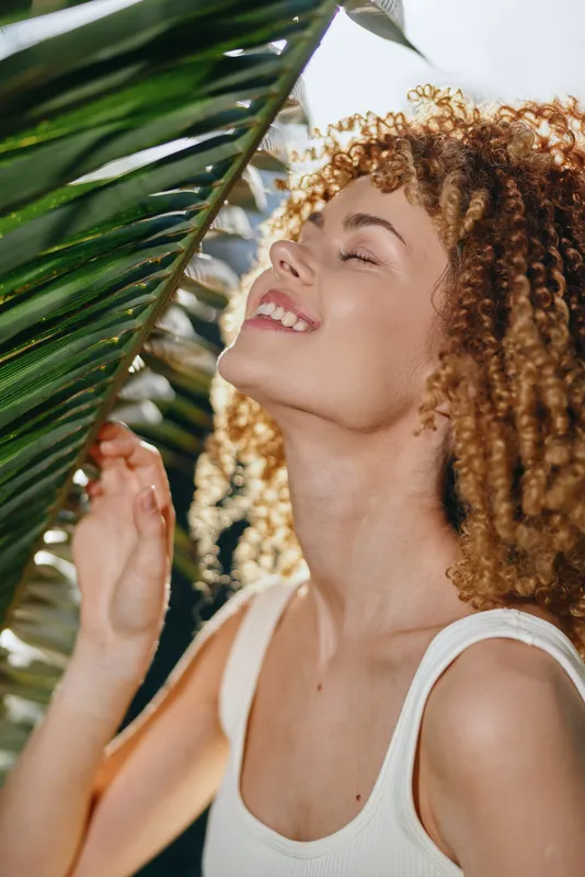 smiling woman with curly hair enjoying sunlight near tropical palm leaves, wearing white tank top in bright natural background, joyful outdoor moment