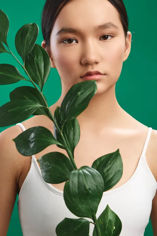 Asian woman stands in a studio, with a large leafy plant framing her calm face. The minimalist setup evokes serenity, nature, and contemporary visual appeal.