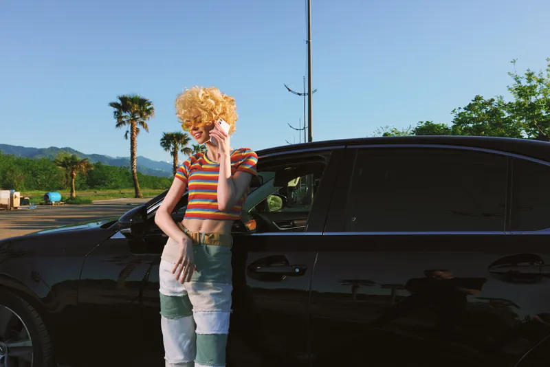 Young woman with curly blond hair stands beside a glossy black car in a sunny outdoor lot, wearing a striped crop top and patchwork jeans, exuding bold retro charm and carefree confidence.