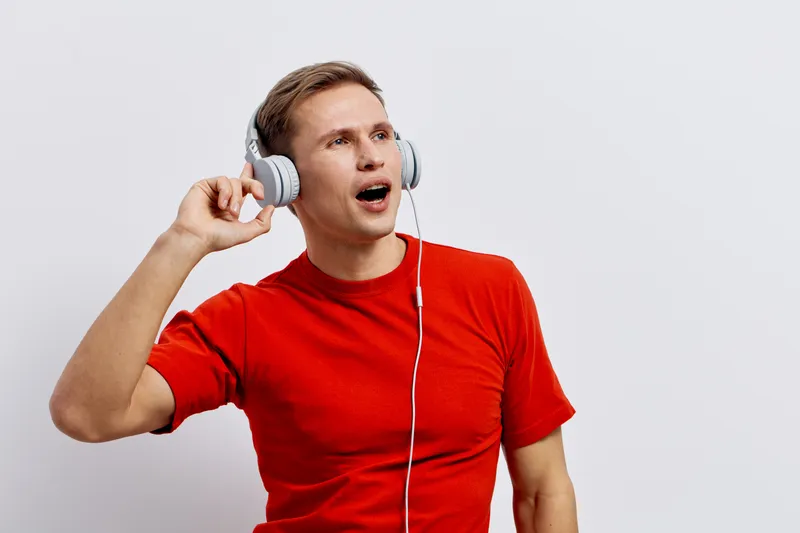 Young man wearing a bright red T-shirt listening to music with headphones, expressing surprise or amazement, standing against a plain light gray background, studio portrait, casual style, lifestyle co