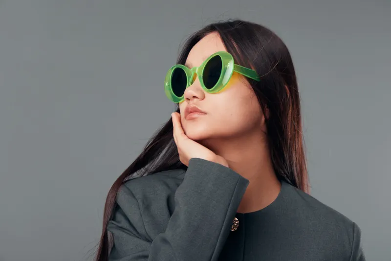 Woman with long dark hair wearing green round sunglasses and gray jacket posing with hand on chin on gray background. Young female model looking sideways with serious expression.