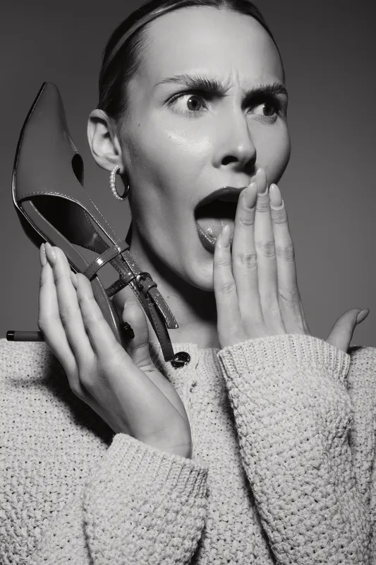 Surprised caucasian woman with light skin holds red high heel shoe near face in studio. Concept for fashion, emotions, and stylish accessories.