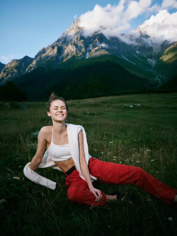 Adventure in a meadow with towering mountains, a smiling woman sits on grass after a workout, bright red pants, white top, towel over shoulder, natural scenery