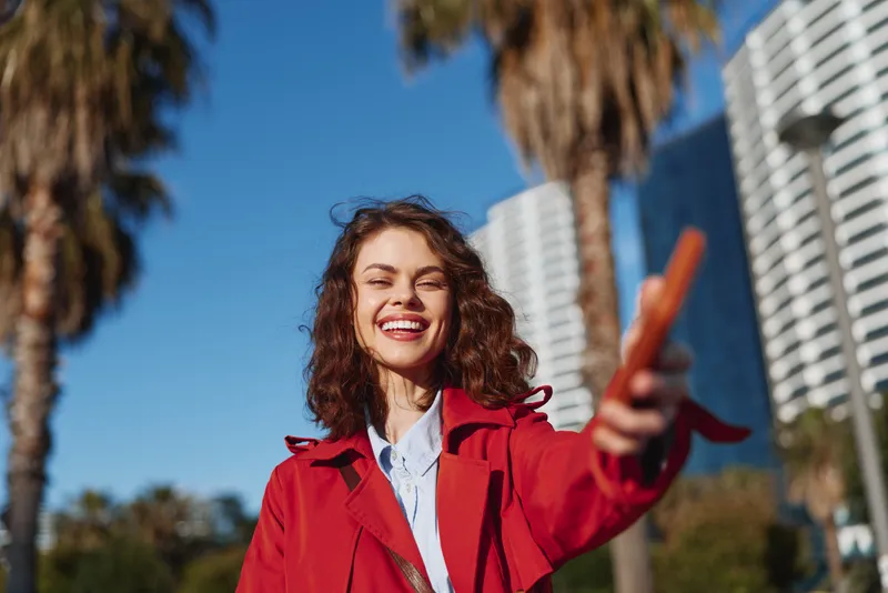 Young brunette woman with curly hair smiles joyfully while pointing forward outdoors on a sunny day near tall buildings and palm trees.