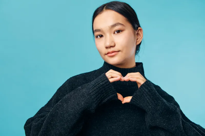 Young woman wearing black sweater making heart shape gesture with hands against blue background. Portrait of smiling female expressing love and positive emotion with casual hairstyle outdoors.