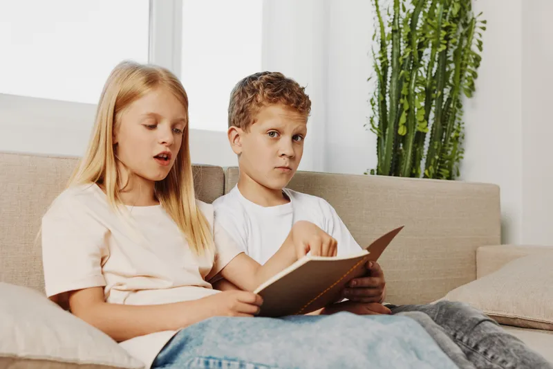 Two children sitting on a beige couch reading a book together in a bright living room with natural light and indoor plants. Concept of childhood learning and sibling bonding.