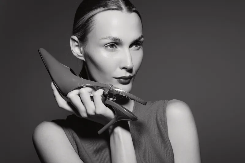 A slender white woman with light skin holds a high heel shoe close to her face, posing thoughtfully in a studio with soft lighting. Concept of fashion and elegance.
