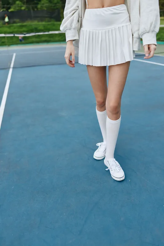 Young woman in a white sporty outfit showcasing confidence on a tennis court, with a pleated skirt and knee high socks, embodying a vibrant athletic lifestyle