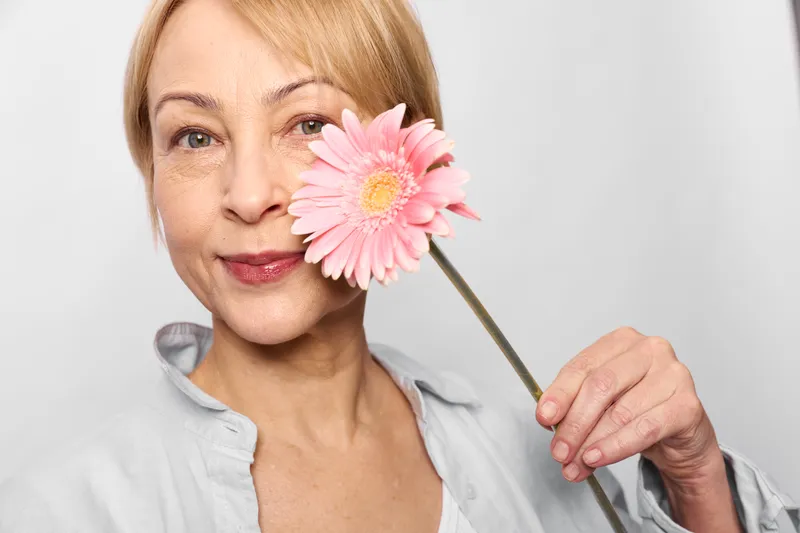 mature woman holding pink flower near cheek, close up portrait in studio, gentle smile, skincare concept, soft light, white background, clean beauty routine