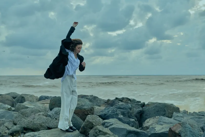 Woman on rocky coast by the sea in a blazer and white trousers, raising a fist in celebration, showcasing movement, outdoors fashion, wind and coastal energy, dramatic mood, bold stance
