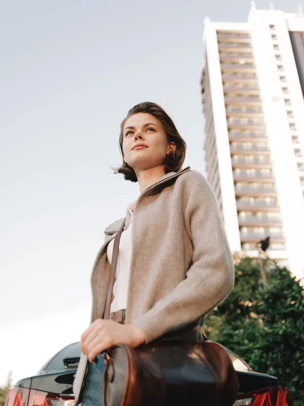 A confident woman stands outdoors in an urban setting, carrying a leather bag and looking upward. The tall buildings and clear sky convey a modern, aspirational lifestyle.