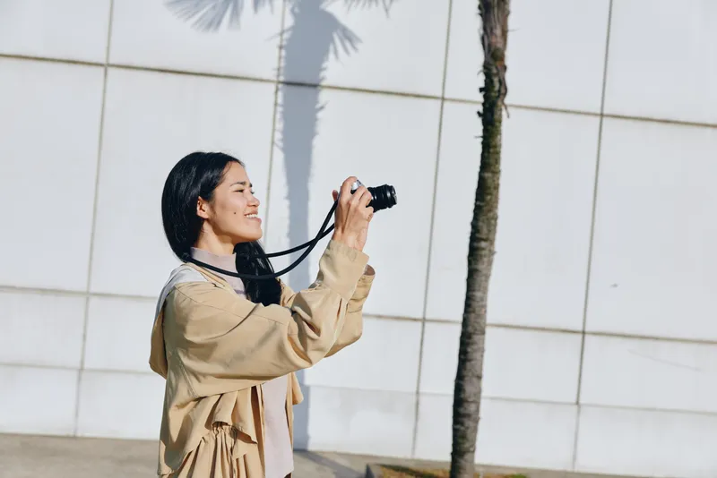 Camera in hand, woman captures outdoor scene under sunny palm shade, a candid street style shoot with lens focused on urban architecture and vibrant daylight