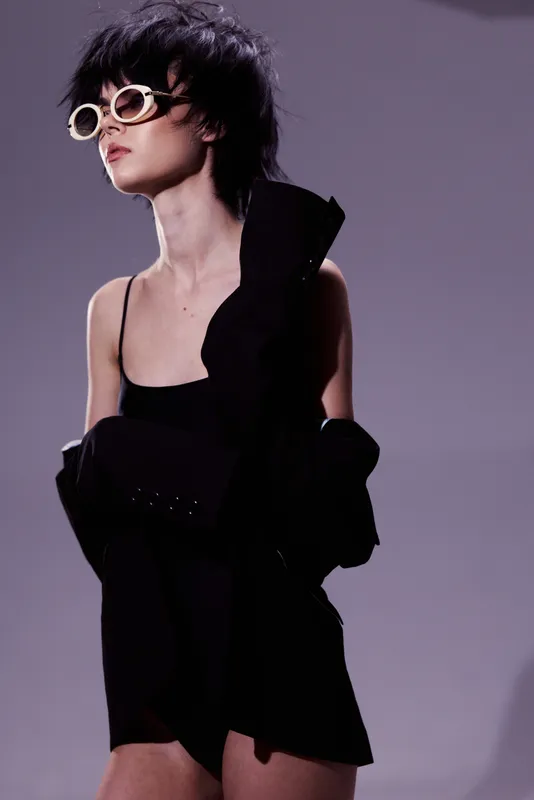 Stylish woman with short dark hair wearing sunglasses and black dress posing confidently in studio lighting.