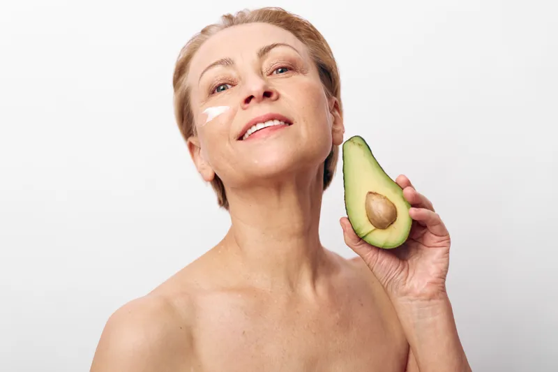 Mature woman with short blonde hair holds half of avocado near face with cream on cheek. Concept of natural skincare, healthy lifestyle, and organic beauty treatments at home.