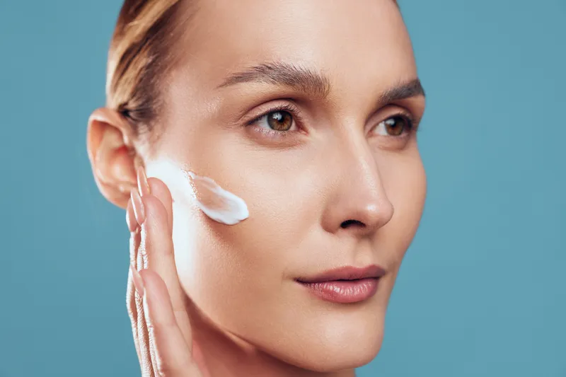 Woman skincare closeup, face cream application on cheek, cleanser foam on skin, beauty spa hygiene concept, serene expression, soft lighting, minimal makeup, turquoise background