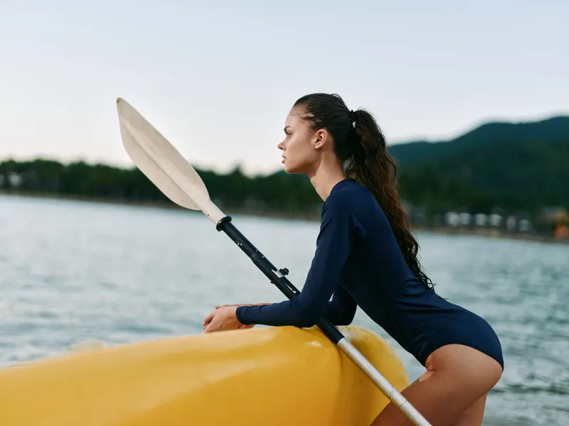 Young woman in a navy wetsuit kneels on a yellow kayak by a calm lake, gazing toward the horizon as the paddle rests, capturing an outdoor adventure moment.