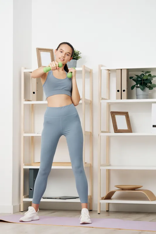 Woman fitness model in blue athletic wear performing shoulder press with dumbbells at home, bright living room setting with plants and shelves, highlighting workout routine and motivation.