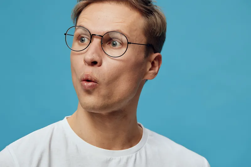 Close-up of surprised young man with glasses and fair skin against blue background, expressing shock or curiosity, casual white t-shirt, studio portrait, people emotion concept