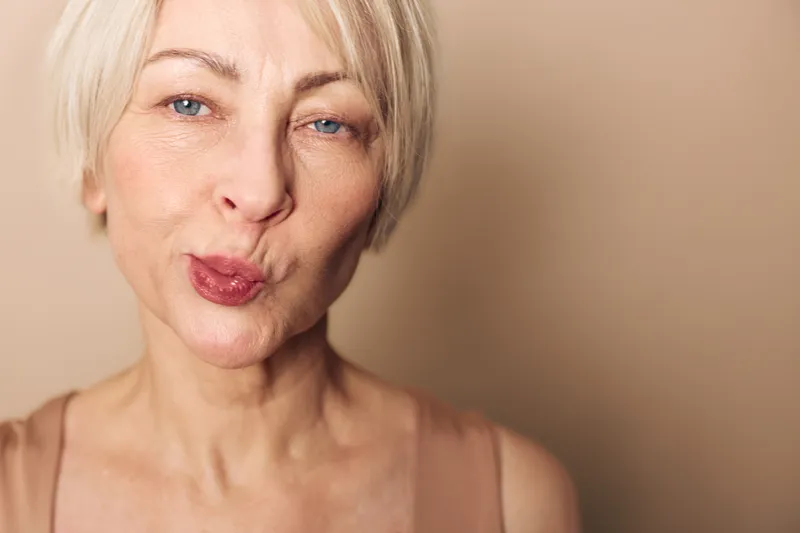 Close-up of mature woman with short blonde hair making playful face, pursing lips, and squinting eyes. Neutral beige background highlights natural skin texture and expression. Concept of joy and