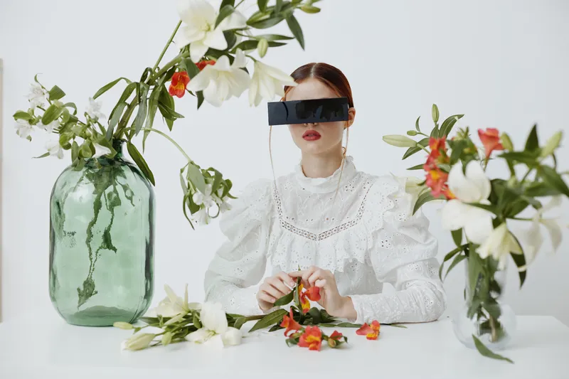 A woman in a vintage white dress wearing futuristic black glasses, surrounded by colorful flowers in vases, symbolizing the fusion of nature and technology
