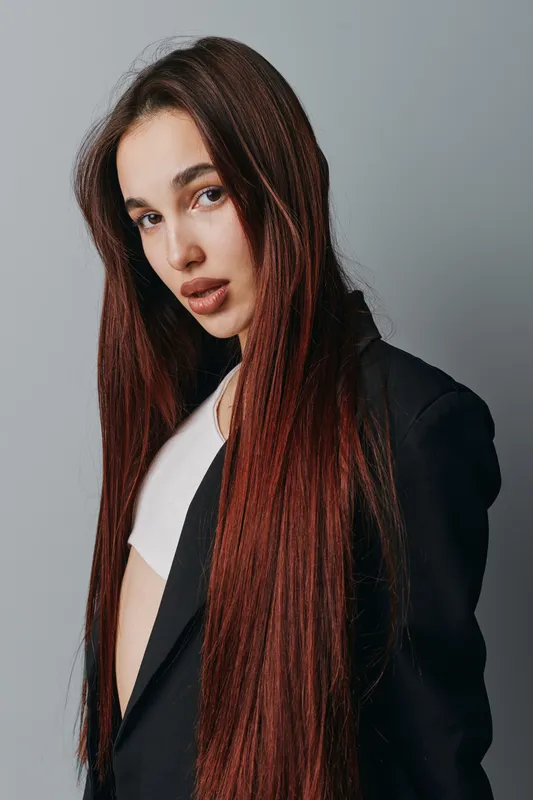 Young woman with long straight hair wearing black blazer and white top posing against gray background. Fashion portrait with natural makeup and confident expression in studio lighting.