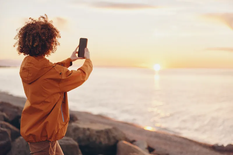 Woman lifestyle at the beach during sunset, curly hair, holding smartphone for photo taking, side view, warm golden light on face, ocean waves behind, travel mood, calm outdoors, peaceful outdoors