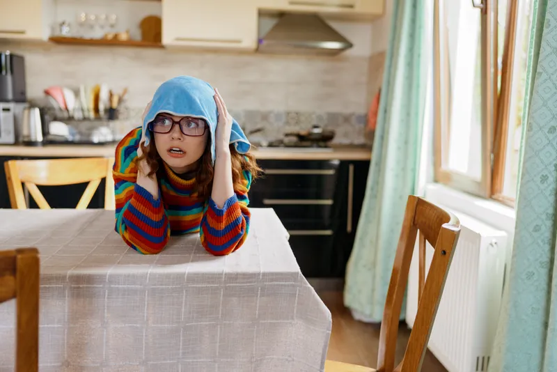 Woman lifestyle home indoors, seated at a dining table in a cozy kitchen, colorful sweater and glasses, relaxed pose, cheerful mood, natural light, everyday moment, warm decor