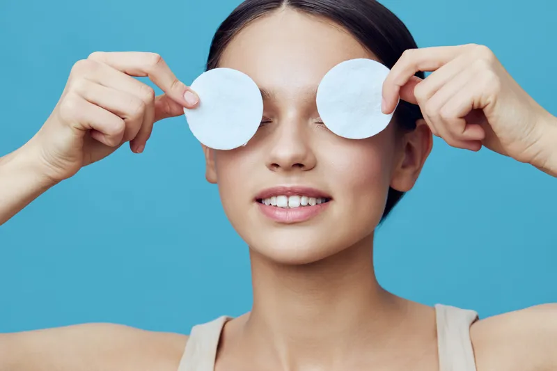 Young woman playfully holding round cotton pads over her eyes, smiling against a blue background, conveying a fun and fresh skincare concept