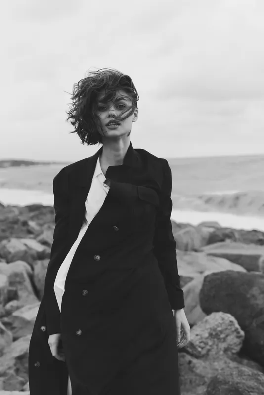Fashion woman on a windy beach wearing a long coat, monochrome scene with sweeping movement, rugged rocks, dramatic coastal atmosphere and outdoor photography