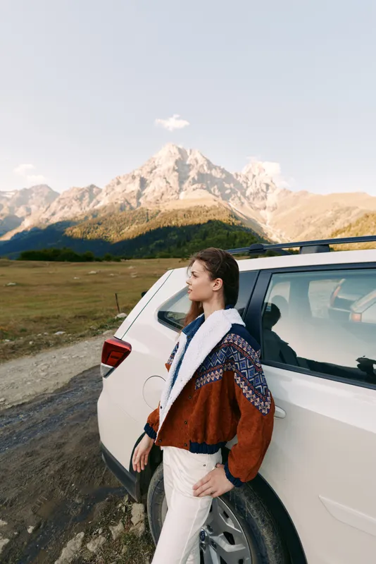Woman, car, mountains, travel, road, nature: Young woman leaning on a white SUV at the roadside with panoramic alpine peaks, cozy jacket, contemplative pose and outdoor adventure mood.