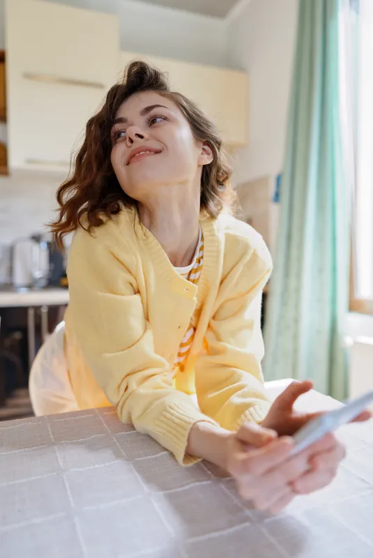 Cheerful woman at home, leaning on a surface, smiling softly, sunlit kitchen behind, casual sweater, warm tones, relaxed mood, cozy atmosphere, everyday lifestyle moment