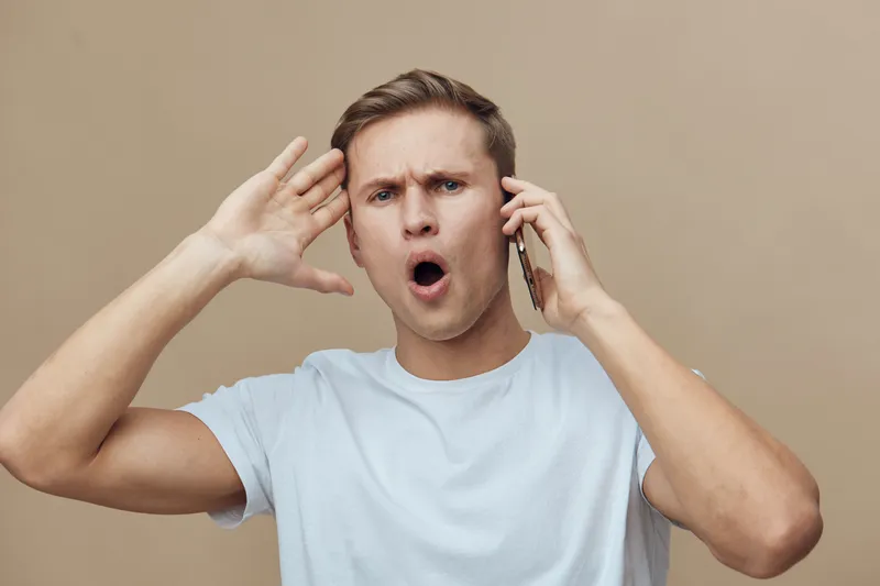 Angry man with short brown hair wearing casual white t-shirt talking on phone expressing frustration and surprise isolated on beige background for emotional, human communication, stress concept