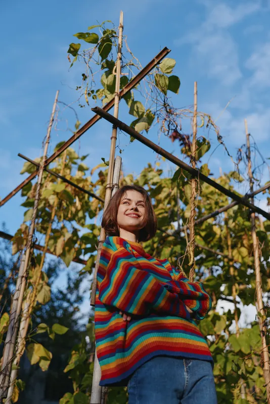 Woman portrait in garden by wooden trellis with climbing plants, smiling in colorful rainbow sweater, casual outdoor lifestyle with natural sunlight and blue sky background.