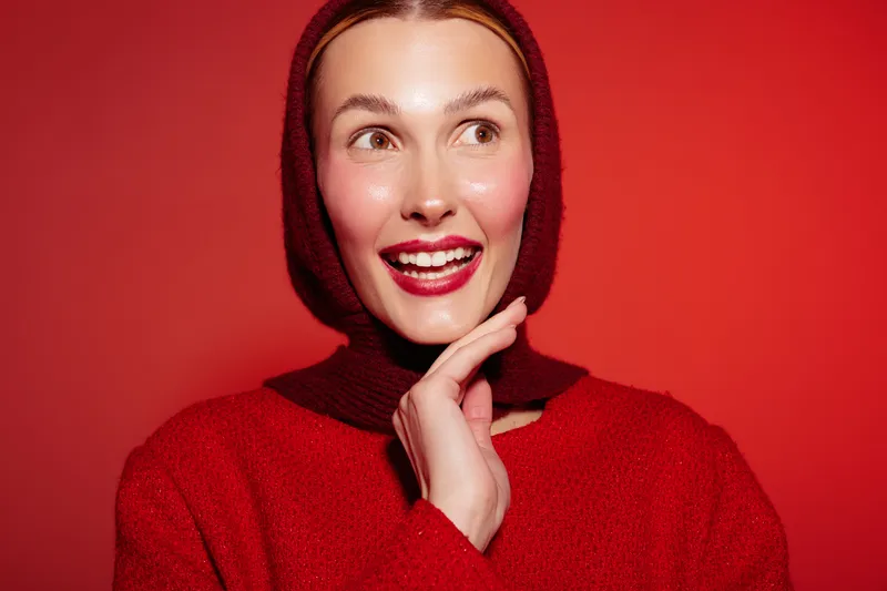Smiling woman in red sweater and hood looks aside with bright lipstick and glowing skin on red background. Concept of fashion, beauty, winter style, and positive emotions.