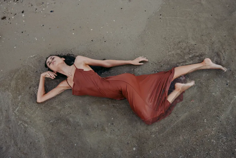 A woman in a flowing rust dress lies on dusty ground, dramatic pose, soft light, and graceful lines creating mood and artistic stillness.