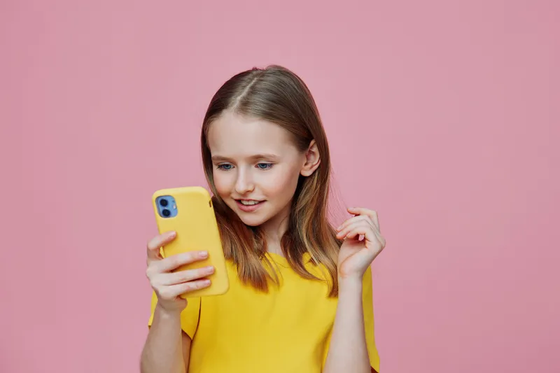A young girl with a cheerful expression, wearing a bright yellow shirt, is engaging with her smartphone against a pink background, showcasing modern technology and youthful enthusiasm