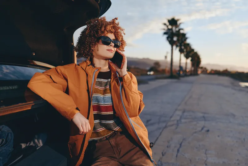 Woman lifestyle curly hair sunglasses phone car sunset fashion street scene, leaning beside a car at dusk holding a phone near her ear, casual jacket, warm light