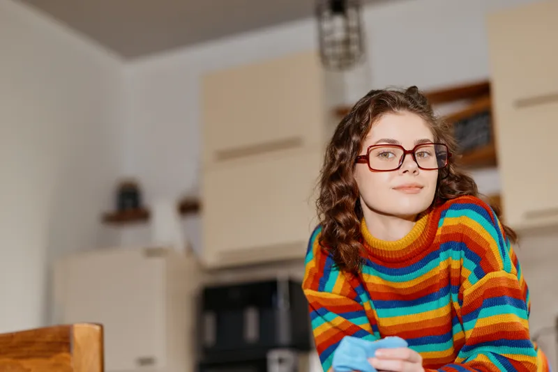 Young woman with curly hair wearing a colorful striped sweater sits indoors in a cozy kitchen, looking at the camera with a gentle smile, creating a warm lifestyle moment of home comfort and casual