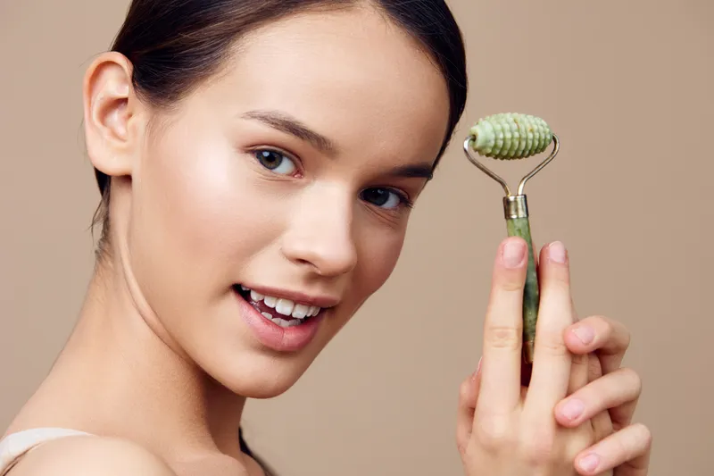 A young woman holding a jade roller, showcasing glowing skin and a joyful expression against a beige background, emphasizing self care and beauty routines