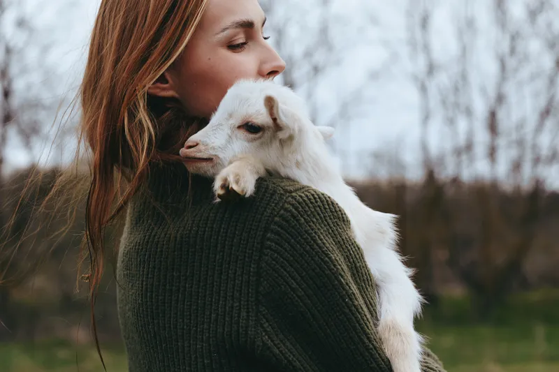 A woman in a dark green sweater holds a young goat close, her face soft with tenderness in a sunny rural setting. A clear lifestyle moment of animal companionship and maternal affection.