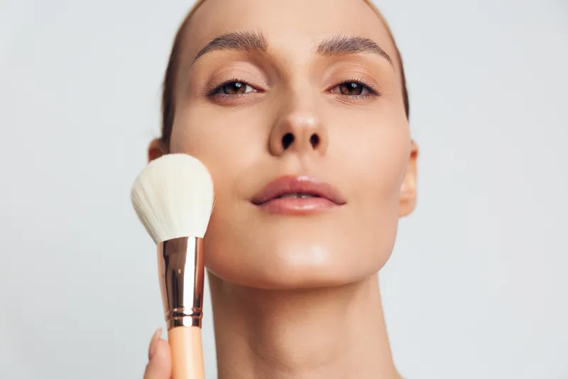 Close-up portrait of a woman applying makeup with a powder brush on a light background. Concept of beauty, skincare, and cosmetic routine with soft natural lighting.
