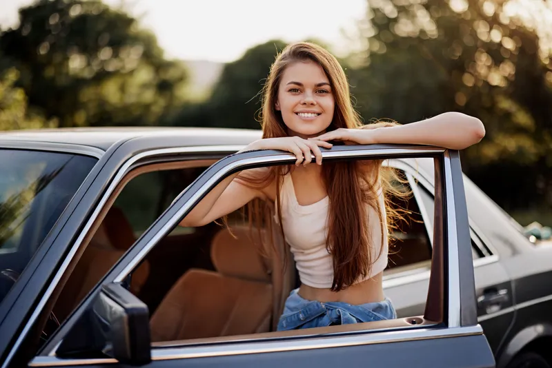 Happy young woman with long hair leaning out of a vintage car, smiling in sunlight surrounded by nature, showcasing a carefree summer vibe.