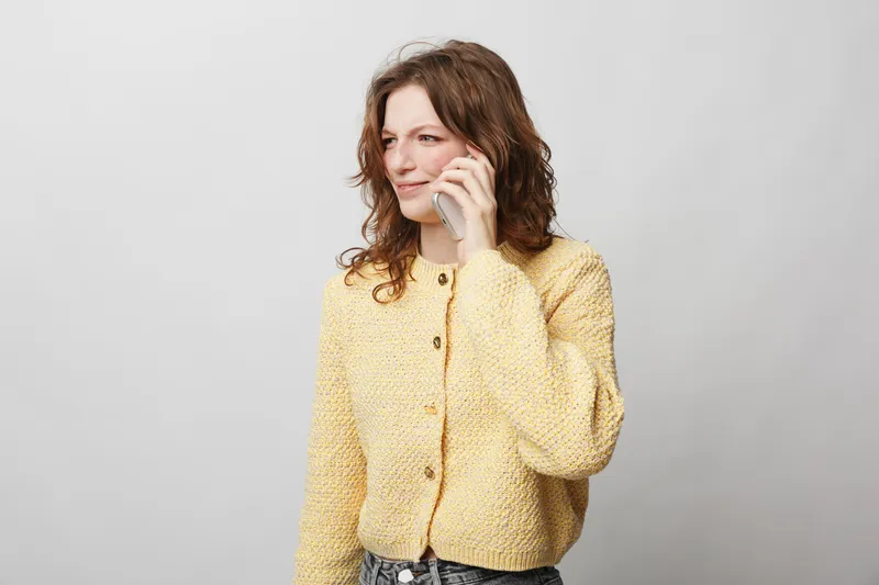 Young woman in yellow knitted sweater talking on smartphone, looking away with slight smile. Casual lifestyle, communication, technology, and positive mood concept on neutral background.