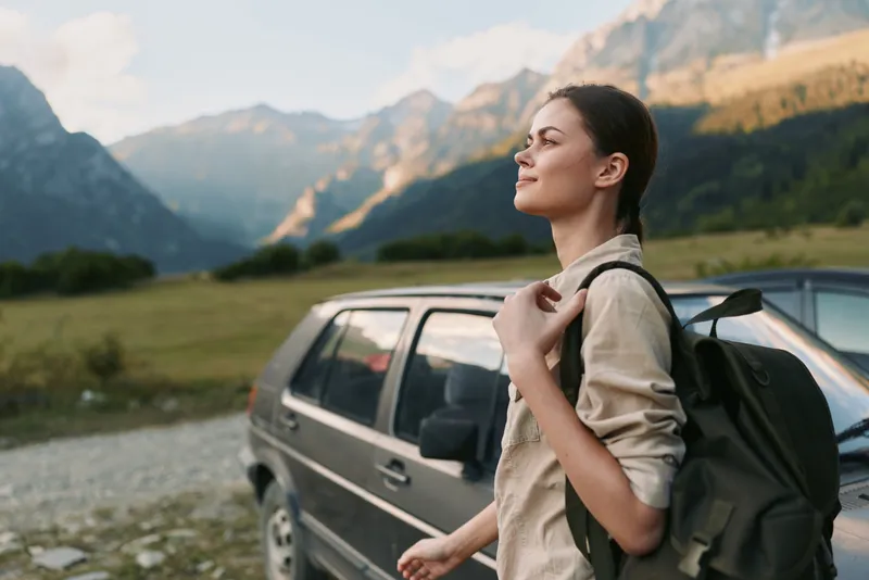 Young woman with a backpack stands near a car in a mountain valley enjoying the fresh air and peaceful nature during a hiking trip.