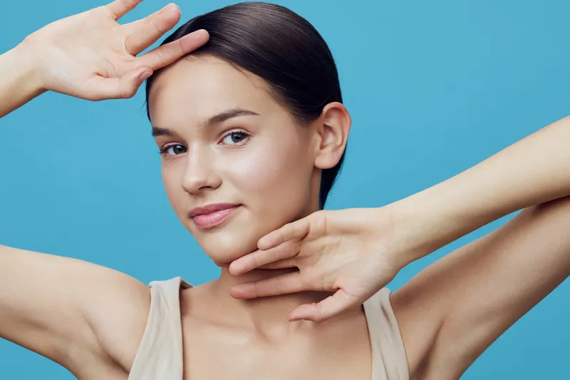 Young woman with natural makeup against a vibrant blue background, showcasing a confident expression and elegant hand poses, perfect for beauty and skincare concepts