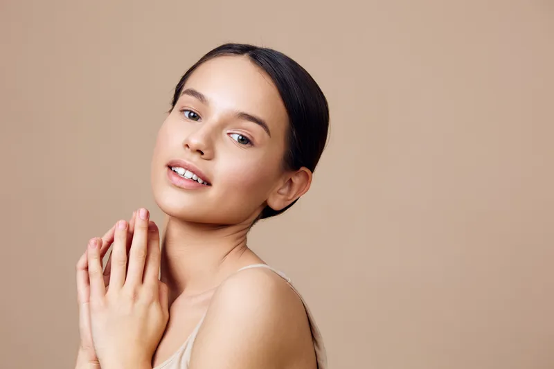 Young woman with smooth skin and a gentle smile, wearing minimal makeup and soft clothing, against a beige background, exuding natural beauty and serenity