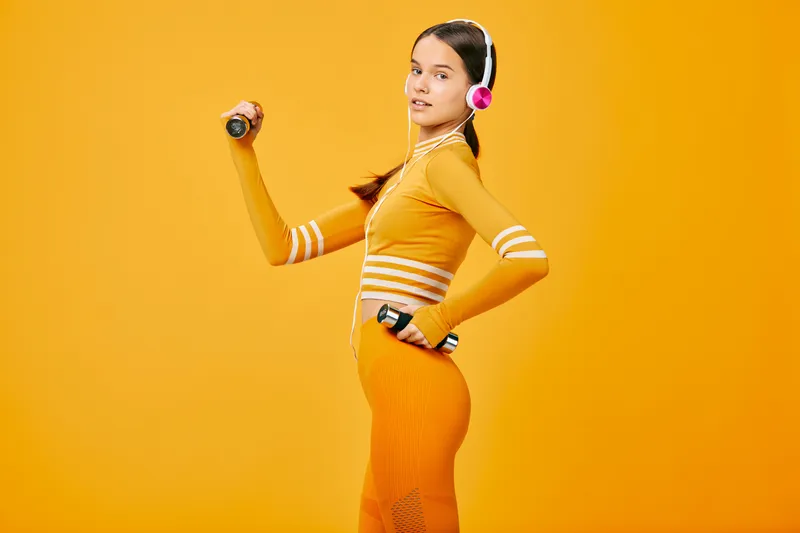 Fitness woman in orange athletic outfit lifts a dumbbell with headphones on, vivid orange studio backdrop, dynamic workout pose, athletic training and motivation for active lifestyle.