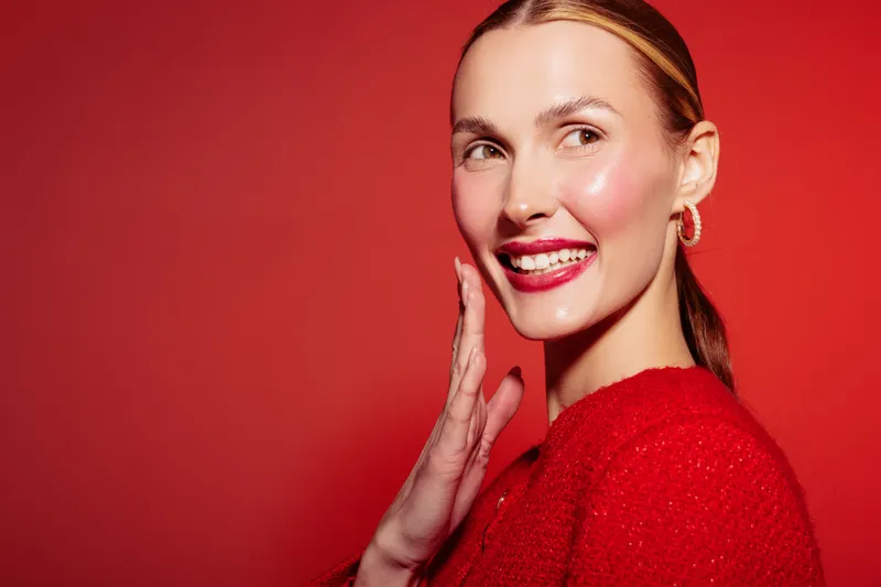 Smiling woman with red lipstick and red sweater poses on red background. Concept of beauty, fashion, makeup, and confident female portrait with bright studio lighting.