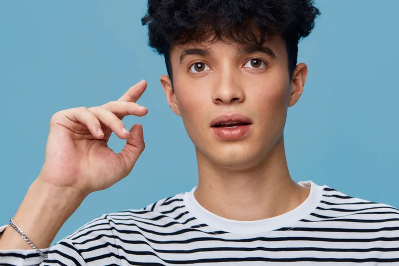 young man with curly hair wearing striped shirt pointing upwards with surprised expression on light blue background, casual style and natural lighting